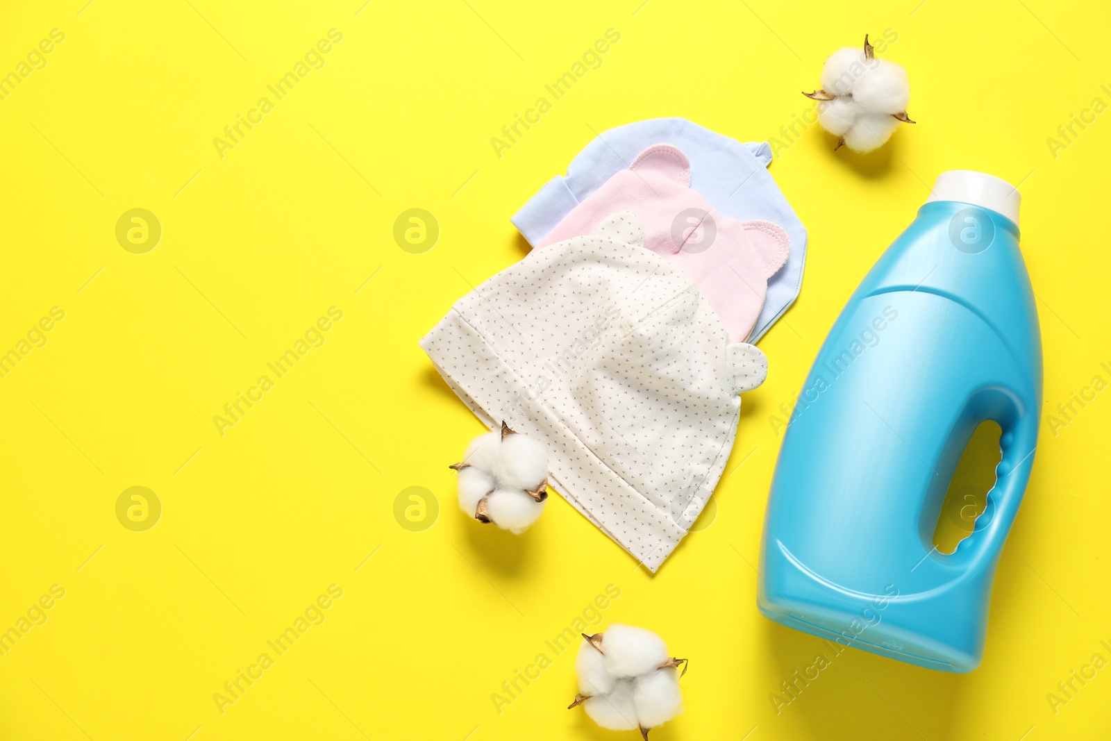 Bottle of fabric softener, cotton flowers and baby hats on yellow background, flat lay. Space for text Photo of Bottle of fabric softener, cotton flowers and baby hats on yellow background, flat lay. Space for text