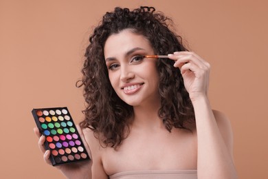 Photo of Smiling woman with eyeshadow palette and brush applying makeup on beige background