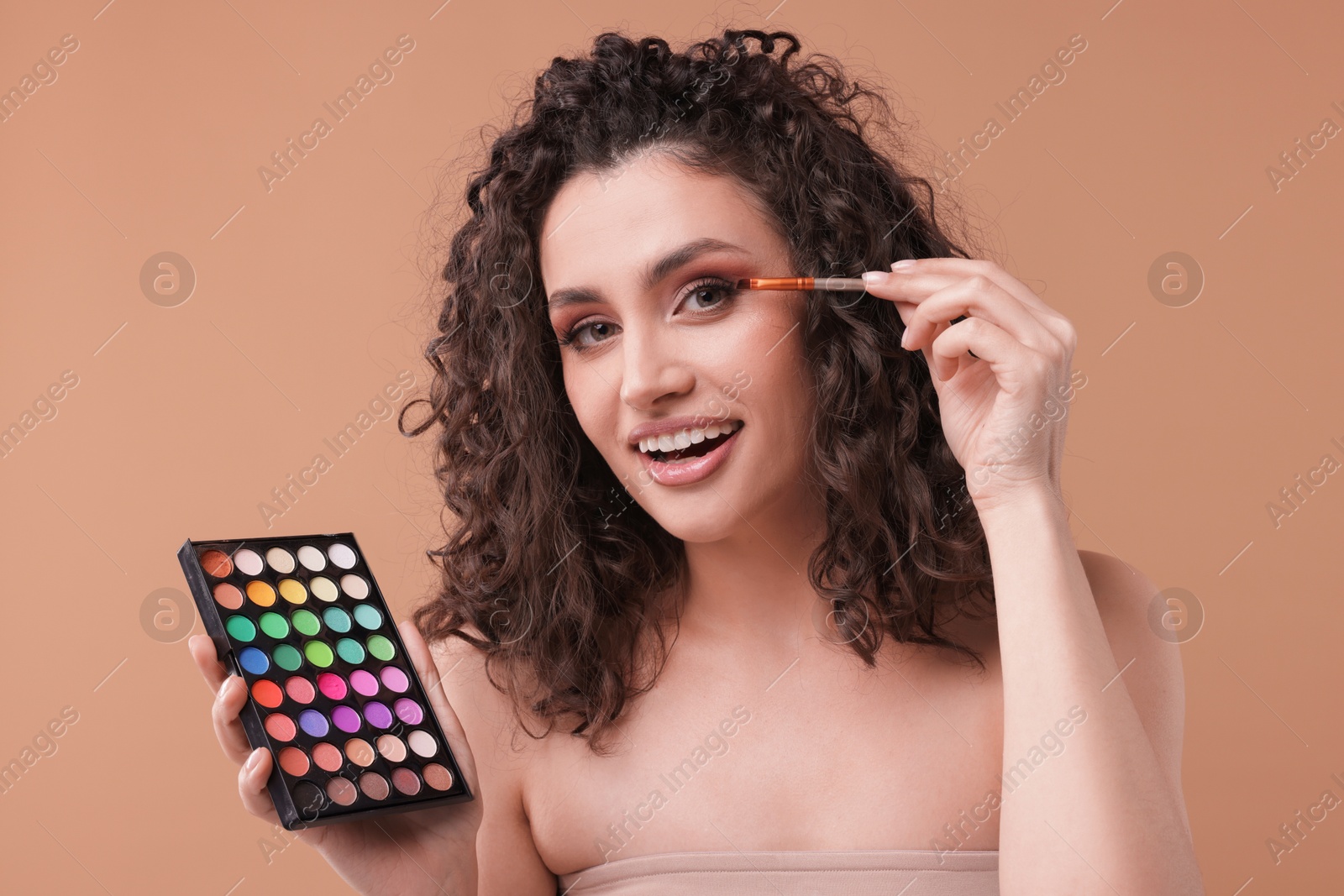 Smiling woman with eyeshadow palette and brush applying makeup on beige background Photo of Smiling woman with eyeshadow palette and brush applying makeup on beige background