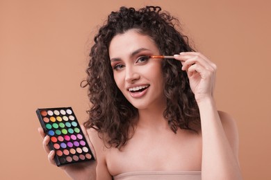 Photo of Smiling woman with eyeshadow palette and brush applying makeup on beige background