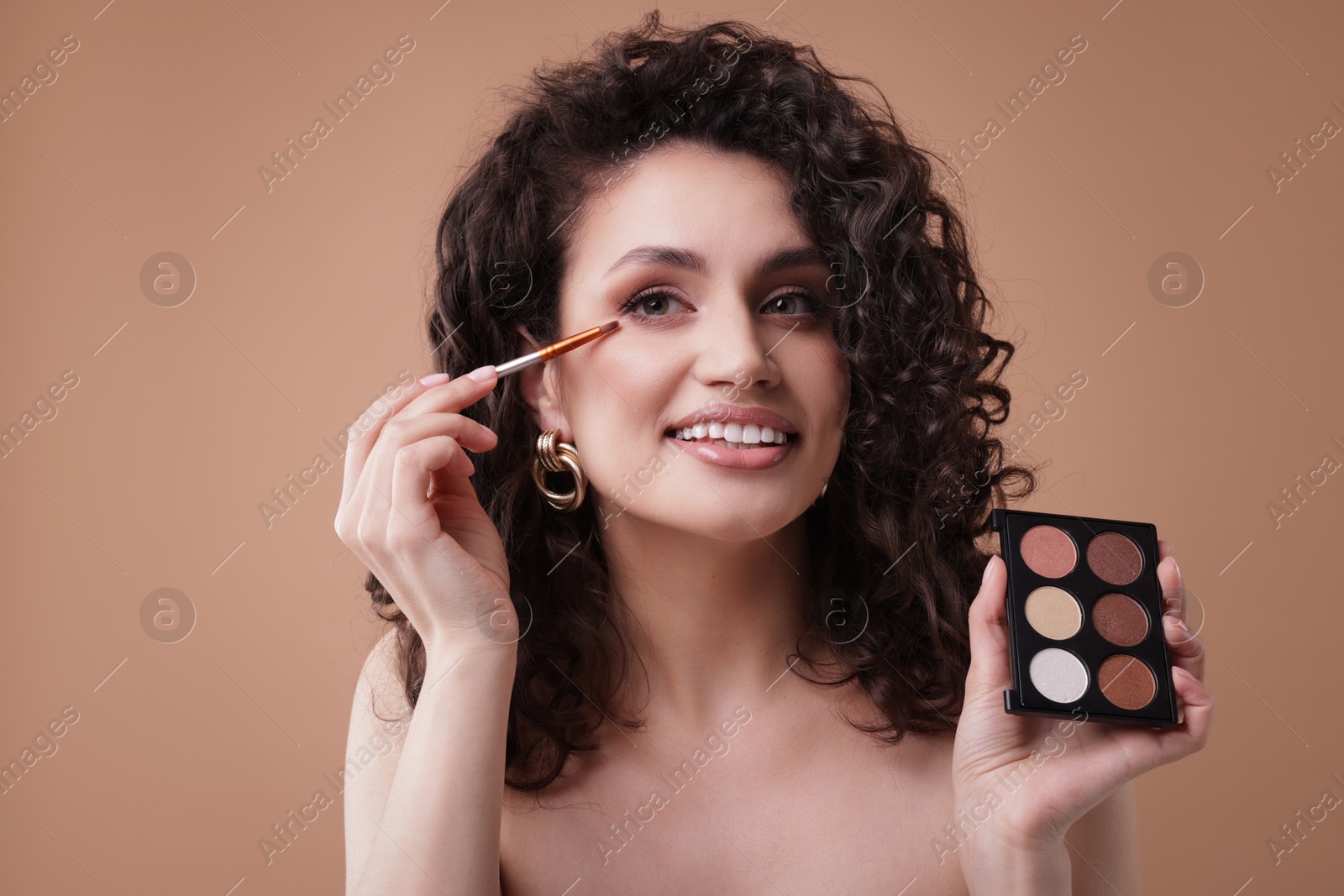 Smiling woman with eyeshadow palette and brush applying makeup on beige background Photo of Smiling woman with eyeshadow palette and brush applying makeup on beige background