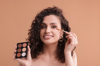 Photo of Smiling woman with eyeshadow palette and brush applying makeup on beige background