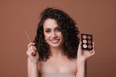 Photo of Smiling woman with eyeshadow palette and brush on brown background
