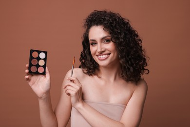 Photo of Smiling woman with eyeshadow palette and brush on brown background