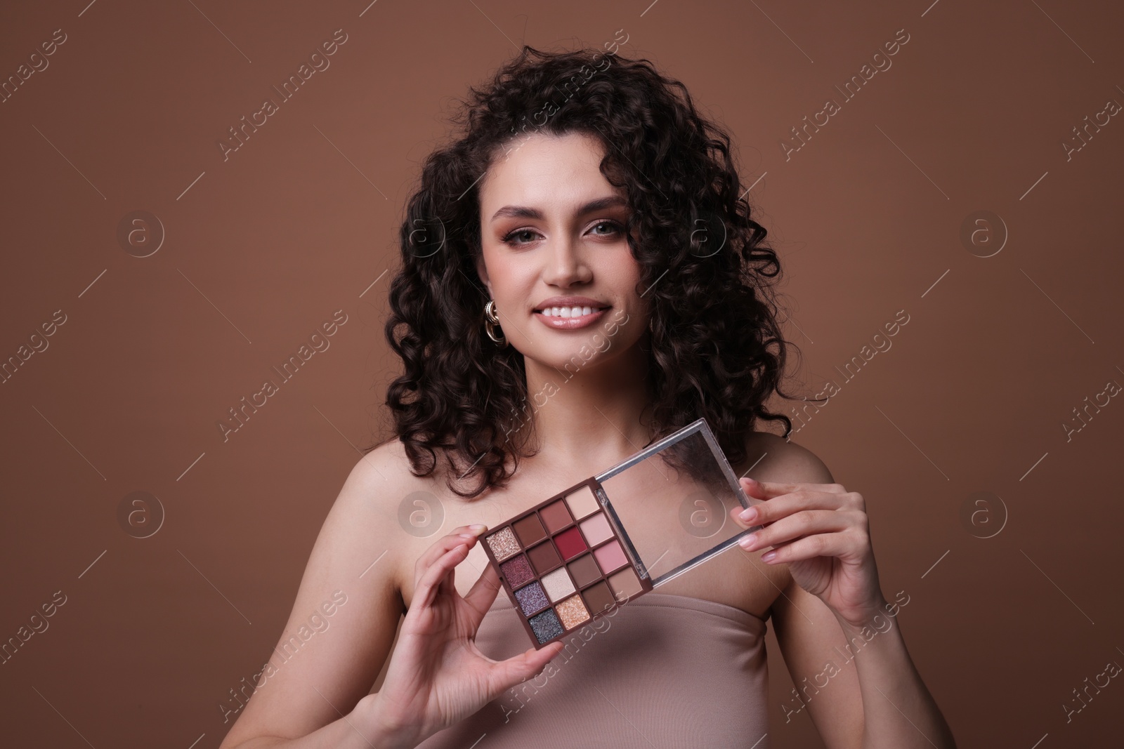 Smiling woman with eyeshadow palette on brown background Photo of Smiling woman with eyeshadow palette on brown background