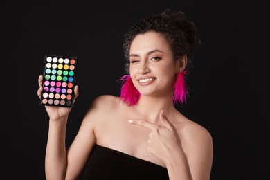 Photo of Smiling woman pointing at bright eyeshadow palette on black background
