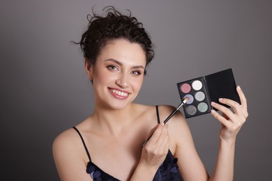 Photo of Smiling woman with eyeshadow palette and brush on grey background