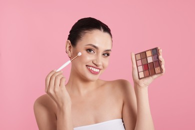 Photo of Smiling woman with eyeshadow palette and brush applying makeup on pink background