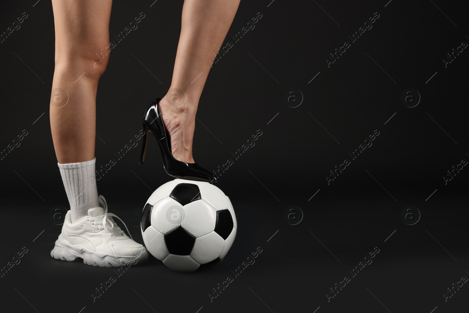 Photo of Woman in high heeled shoe and sneaker with soccer ball on dark background, closeup. Space for text