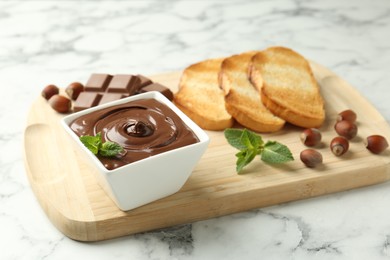 Photo of Tasty chocolate spread, pieces, toasts, mint and hazelnuts on white marble table, closeup