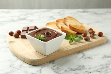 Photo of Tasty chocolate spread, pieces, toasts, mint and hazelnuts on white marble table, closeup