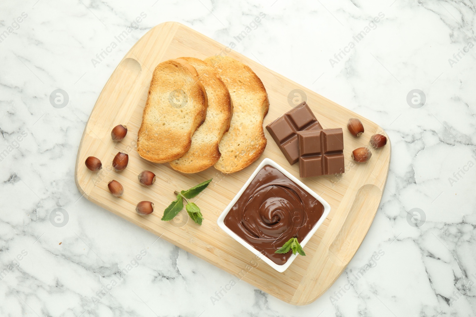 Photo of Tasty chocolate spread, pieces, toasts, mint and hazelnuts on white marble table, top view