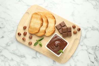 Photo of Tasty chocolate spread, pieces, toasts, mint and hazelnuts on white marble table, top view