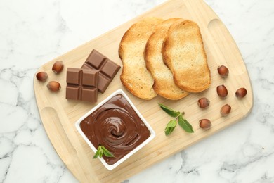 Photo of Tasty chocolate spread, pieces, toasts, mint and hazelnuts on white marble table, top view