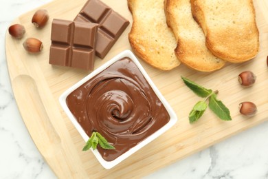Photo of Tasty chocolate spread, pieces, toasts, mint and hazelnuts on white marble table, top view
