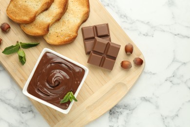 Photo of Tasty chocolate spread, pieces, toasts, mint and hazelnuts on white marble table, top view