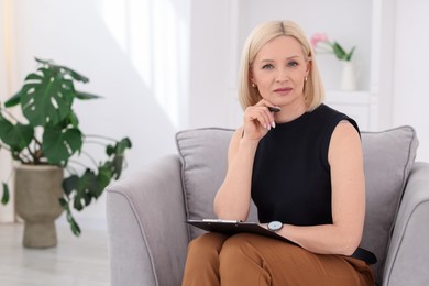 Photo of Professional psychologist with clipboard and pen in armchair indoors