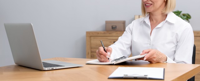 Photo of Professional psychologist taking notes while working with laptop at table in office, closeup