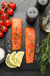Photo of Seafood. Pieces of salmon fillet, spices, tomatoes, oil and lemon slices on black textured table, flat lay