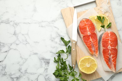 Photo of Seafood. Fresh raw salmon steaks, parsley, pieces of lemon and knife on white marble table, flat lay. Space for text