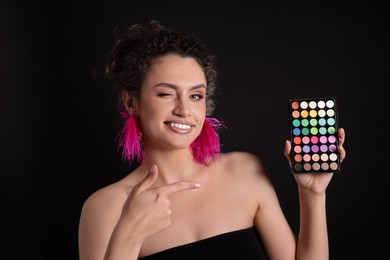 Photo of Smiling woman pointing at bright eyeshadow palette on black background