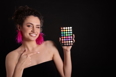 Photo of Smiling woman pointing at bright eyeshadow palette on black background. Space for text