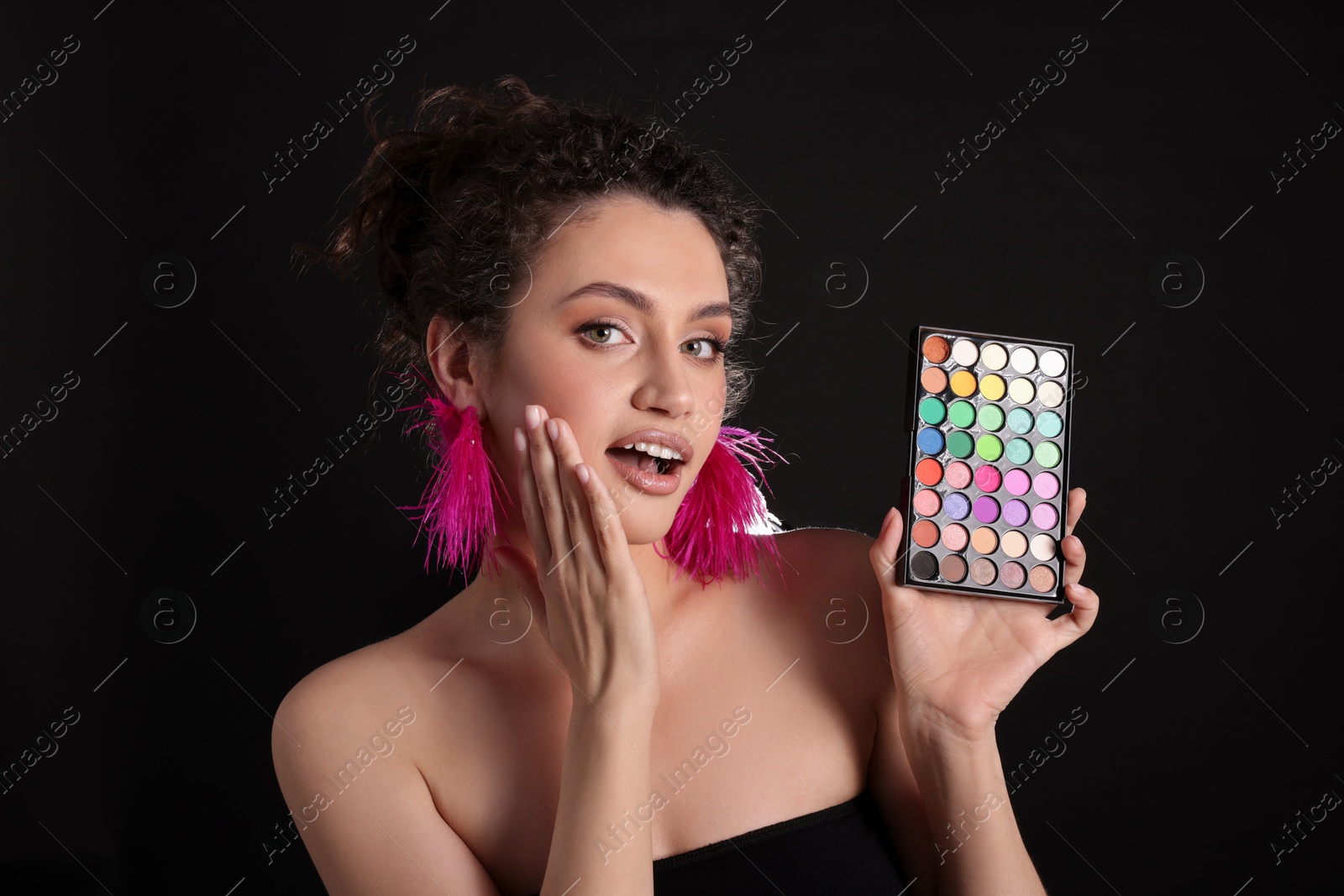 Surprised woman with bright eyeshadow palette on black background Photo of Surprised woman with bright eyeshadow palette on black background