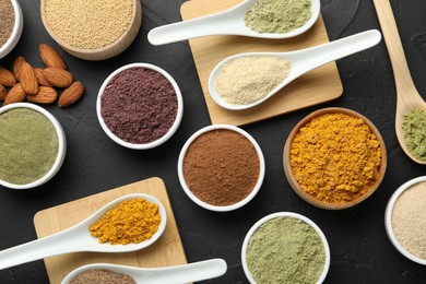 Photo of Different superfood powders in spoons and bowls on dark textured table, flat lay