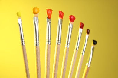 Photo of Many brushes with color paints on yellow background, closeup