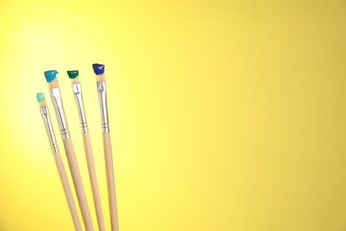 Photo of Many brushes with color paints on yellow background, closeup. Space for text