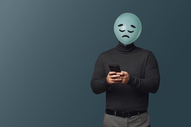 Image of Man with smartphone hiding behind balloon with sad face on dark grey background. Space for text