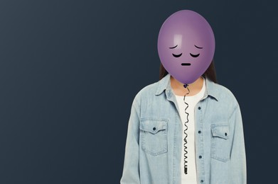 Image of Woman hiding behind violet balloon with sad face on dark grey background. Space for text