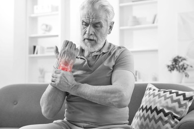 Image of Arthritis. Senior man suffering from wrist pain on sofa at home. Black and white toning with red color accent