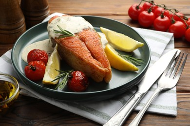 Photo of Seafood. Tasty salmon steak served with sauce, tomatoes, rosemary and lemon on wooden table, closeup