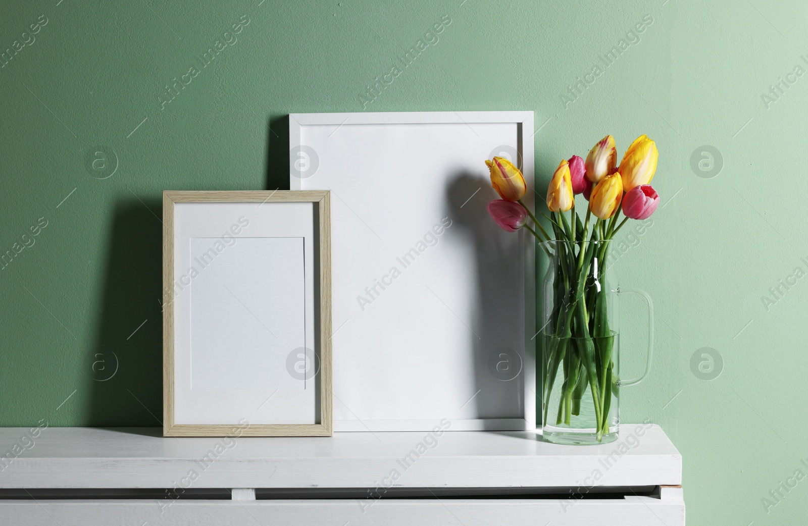 Empty picture frames and beautiful tulips on white shelf near pale green wall indoors Photo of Empty picture frames and beautiful tulips on white shelf near pale green wall indoors