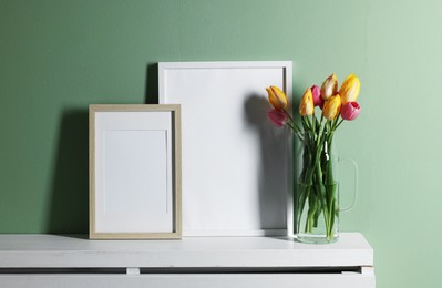 Photo of Empty picture frames and beautiful tulips on white shelf near pale green wall indoors