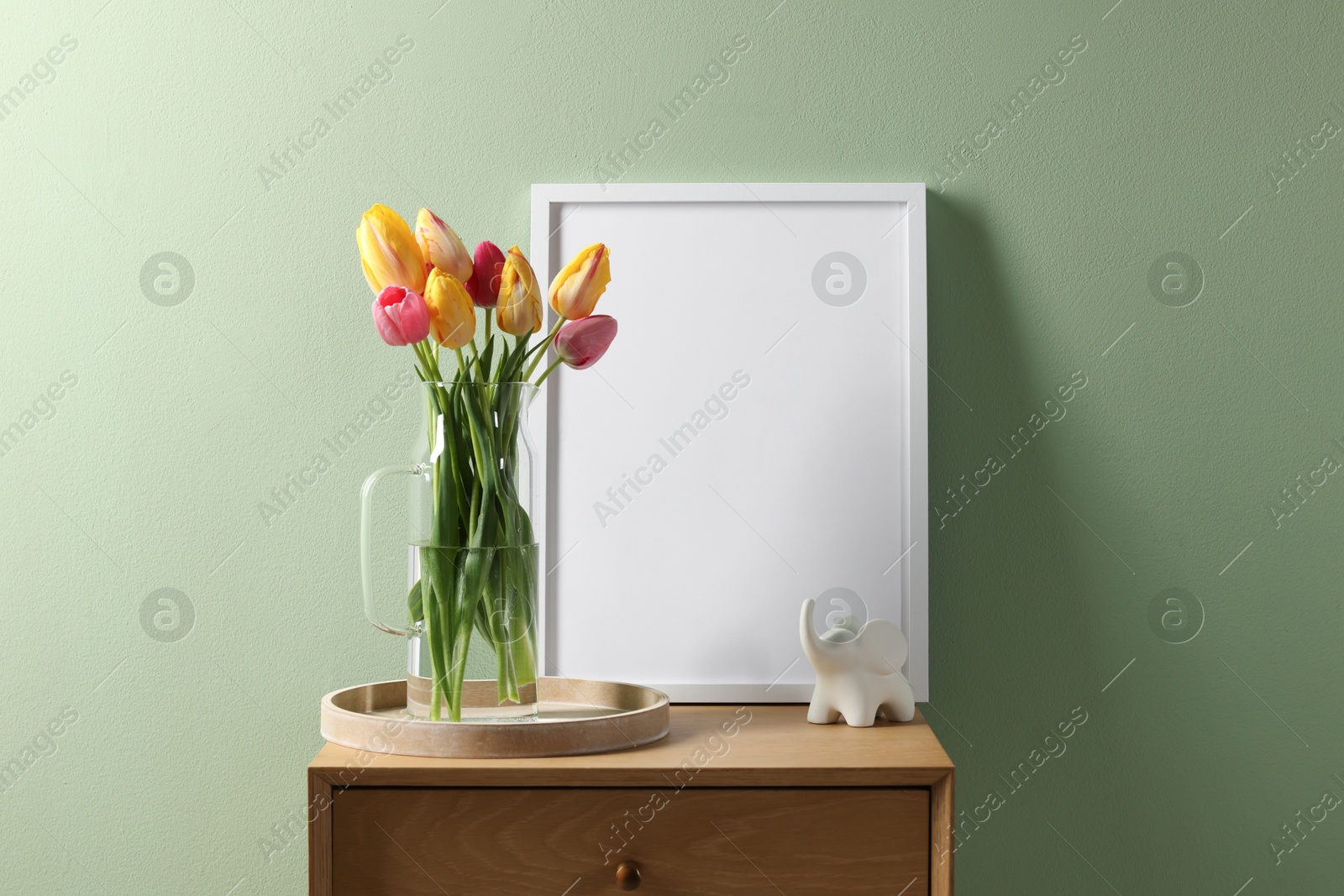 Empty picture frame, decor and beautiful tulips on bedside table near pale green wall indoors Photo of Empty picture frame, decor and beautiful tulips on bedside table near pale green wall indoors