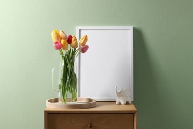 Photo of Empty picture frame, decor and beautiful tulips on bedside table near pale green wall indoors