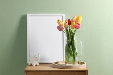 Photo of Empty picture frame, decor and beautiful tulips on bedside table near pale green wall indoors