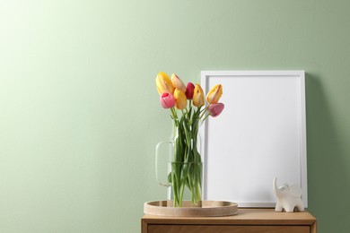 Photo of Empty picture frame, decor and beautiful tulips on bedside table near pale green wall indoors, space for text