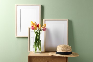 Photo of Empty picture frames, straw hat and beautiful tulips near pale green wall indoors