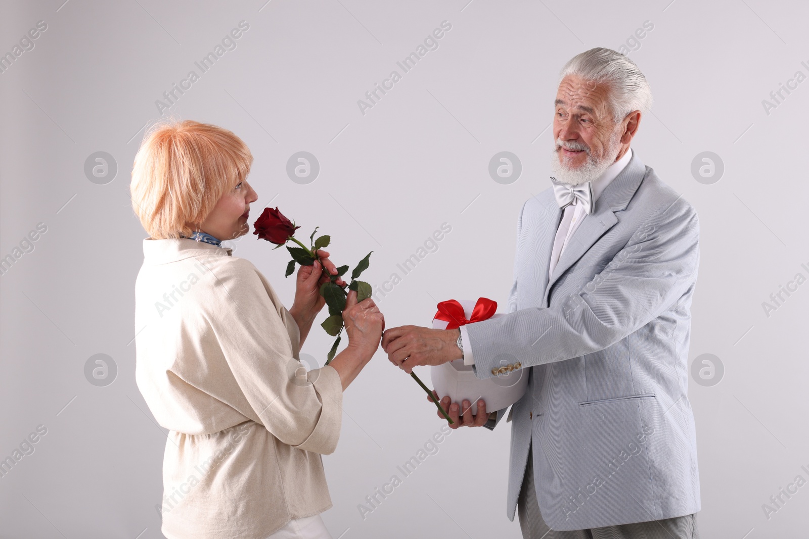 Lovely senior couple with gift box and rose on light grey background Photo of Lovely senior couple with gift box and rose on light grey background