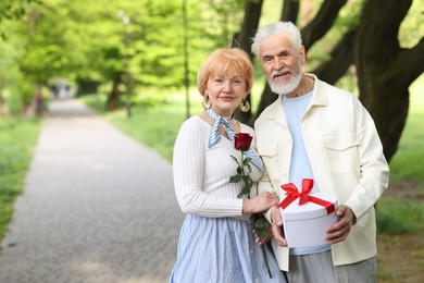 Photo of Lovely senior couple with gift box and rose in park, space for text