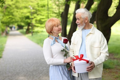 Photo of Lovely senior couple with gift box and rose in park, space for text