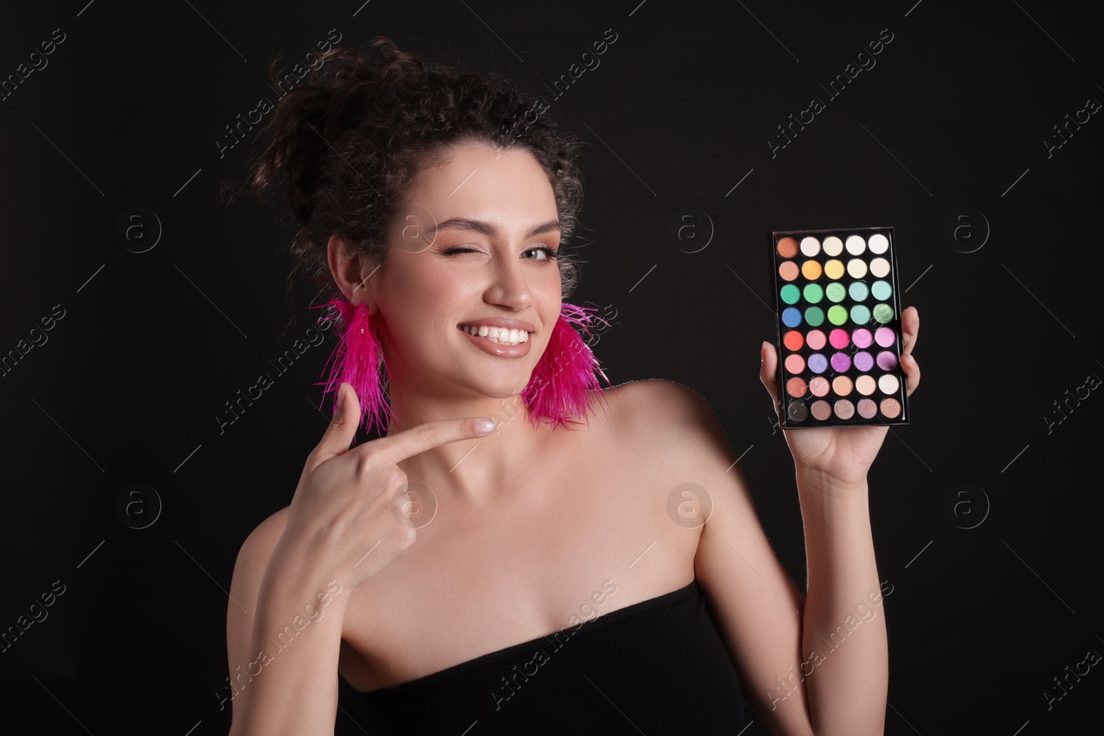 Smiling woman pointing at bright eyeshadow palette on black background Photo of Smiling woman pointing at bright eyeshadow palette on black background
