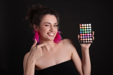 Photo of Smiling woman pointing at bright eyeshadow palette on black background