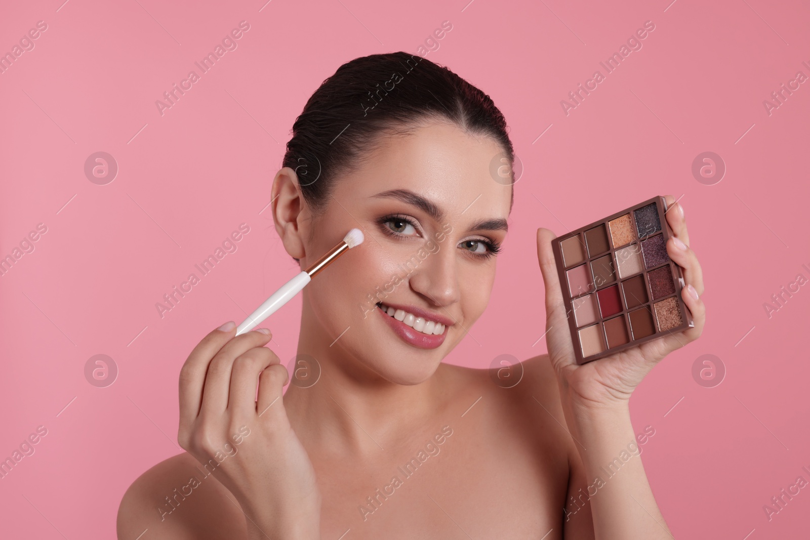 Smiling woman with eyeshadow palette and brush applying makeup on pink background Photo of Smiling woman with eyeshadow palette and brush applying makeup on pink background