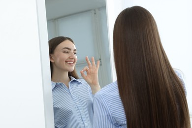 Photo of Beautiful woman showing ok gesture near mirror indoors