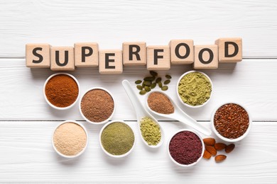 Photo of Different superfoods and word made of cubes on white wooden table, flat lay