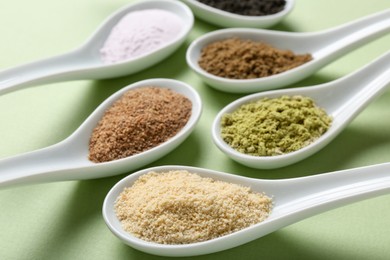 Photo of Different superfood powders in spoons on pale green background, closeup